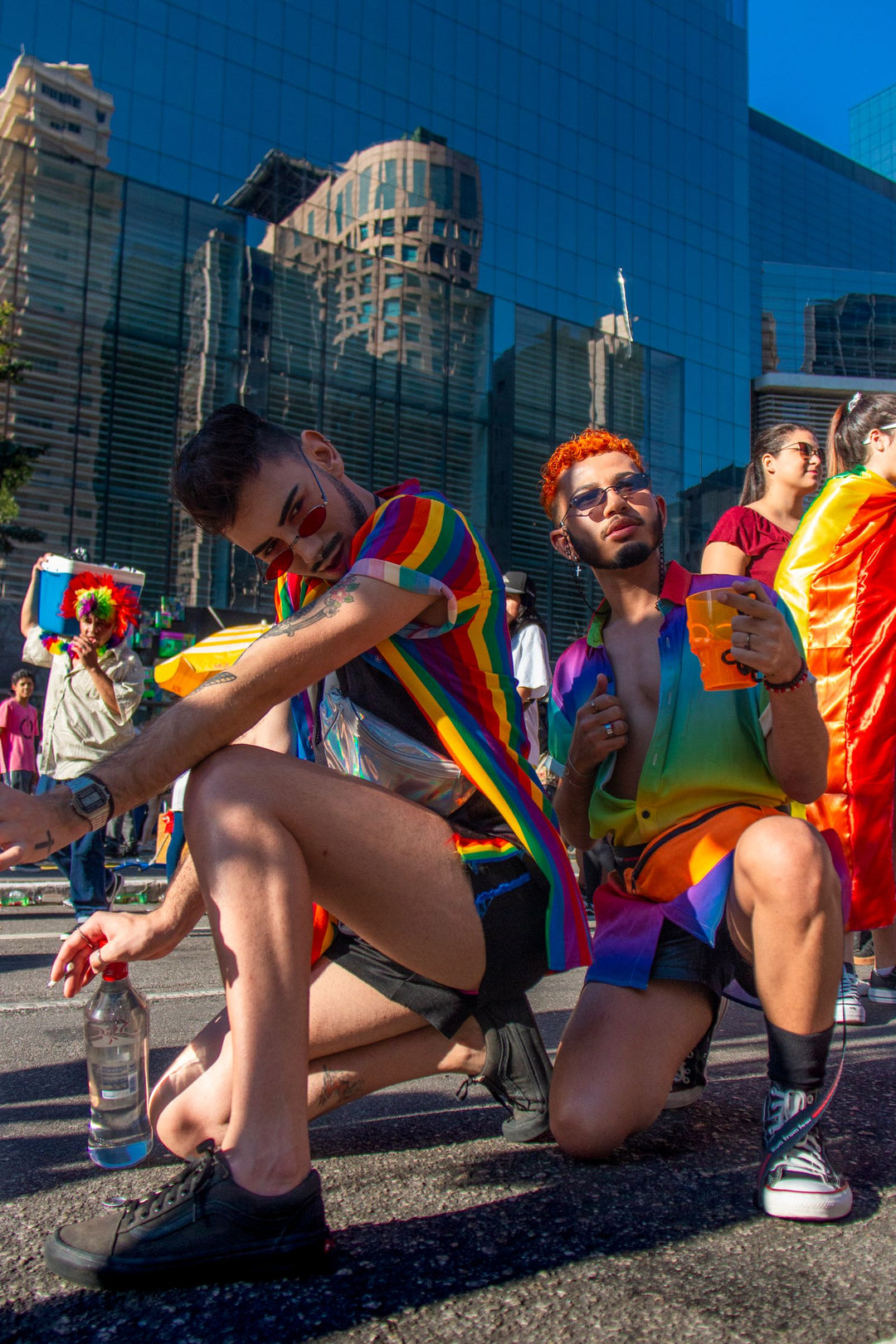 Two men in rainbow tops and shorts posing at a Pride parade, showing men's Pride outfit ideas