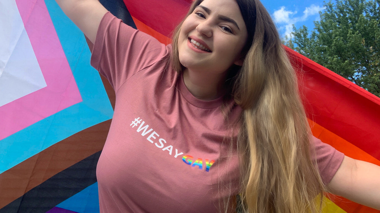 Woman with #wesaygay shirt in mauve with progressive pride flag behind her